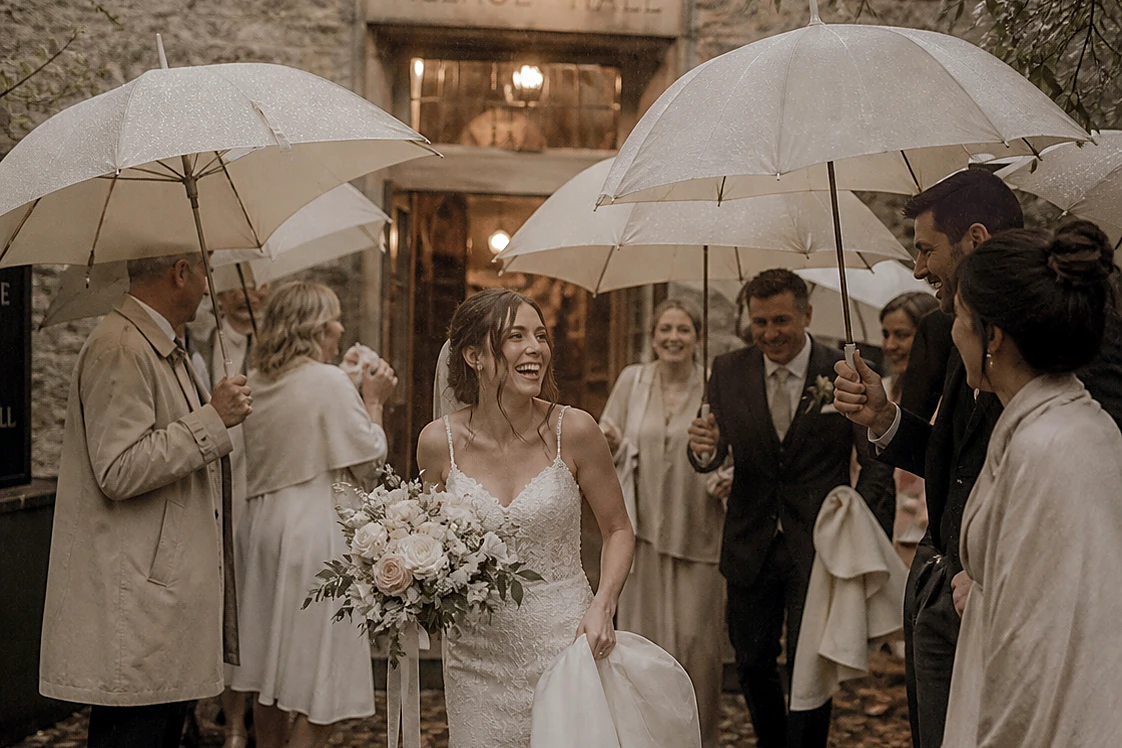 Guests sheltering under cream umbrellas outside a village hall in the rain