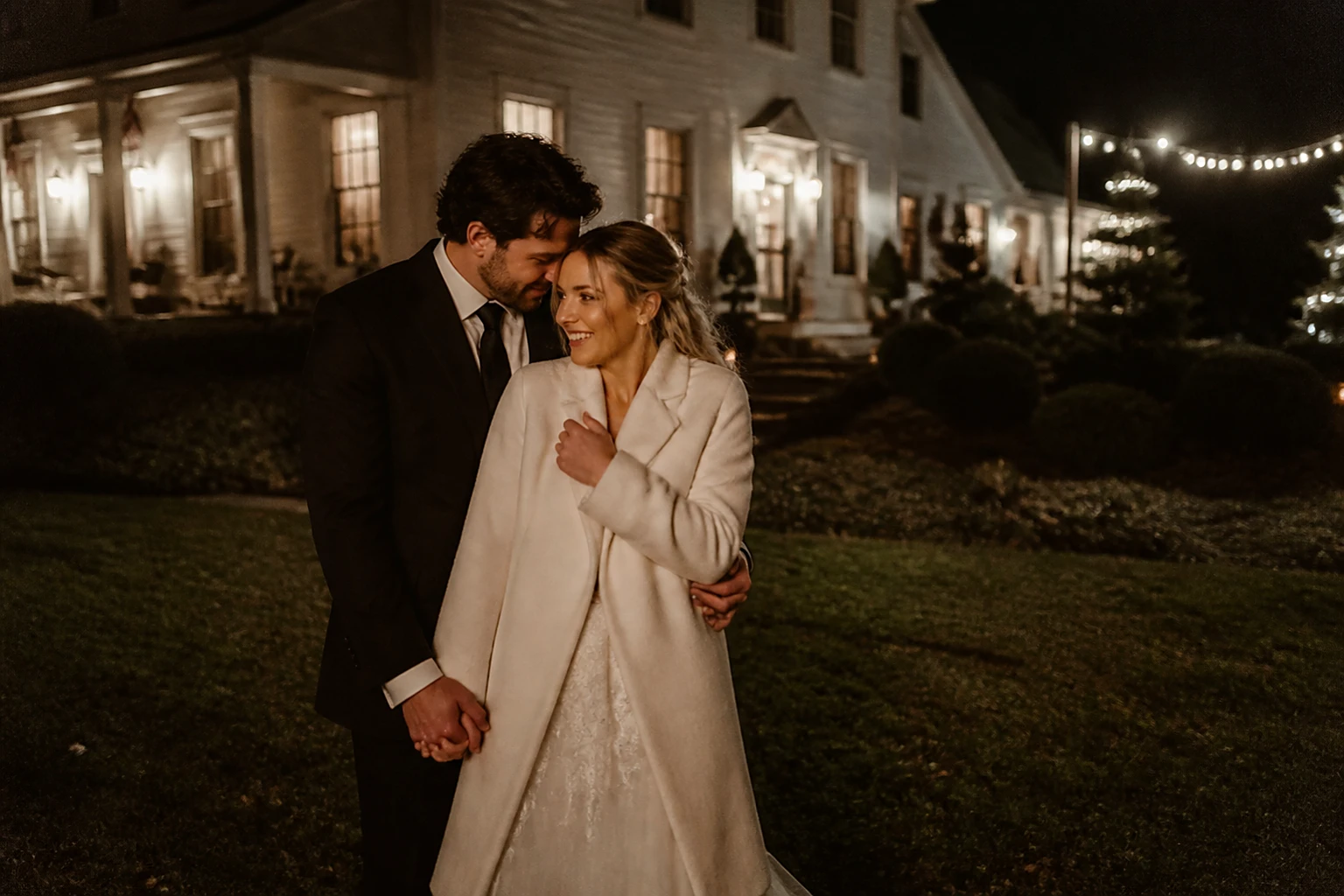 A night portrait of a couple outside a lit farmhouse under string lights